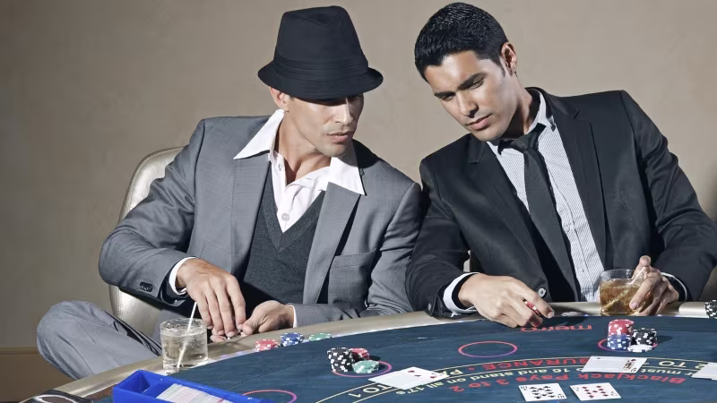 Two people in suits sitting at a casino table with blackjack chips, playing cards, and drinks.